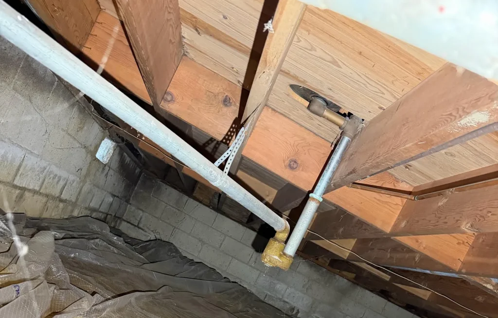 A dirty crawl space showing exposed wooden joists above a brick foundation wall. A white pipe runs horizontally along the wall, connecting to a vertical pipe with yellow insulation. The ground is covered by a dirty, translucent vapor barrier.